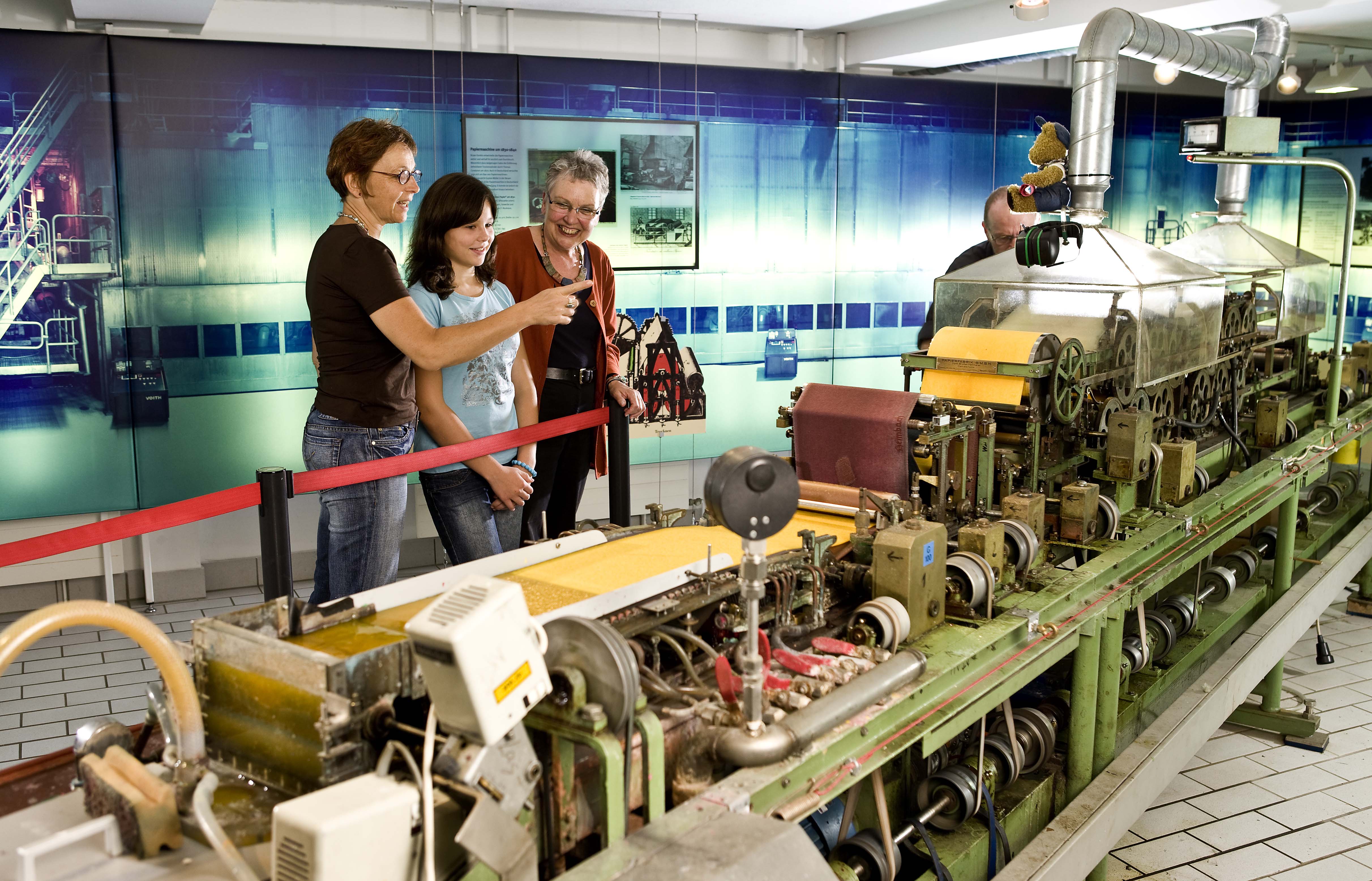 Drei Frauen stehen vor einer industriellen Papiermaschine, die sich im LVR-Industriemuseum Alte Dombach befindet